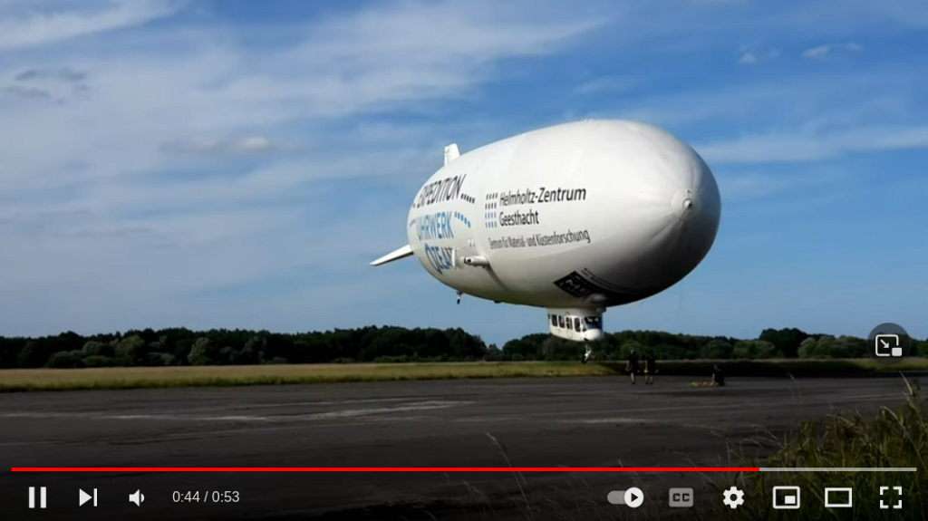 Videovorschau. Ein weißer Zeppelin an einer Landebahn. Auf dieser stehen Menschen. 